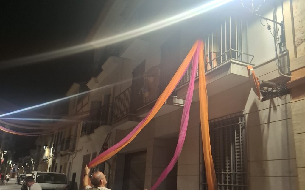 A LES PORTES DE LA FESTA, ADORNANT CARRER. ENTRE TELES I POMPONS, EL CARRER SIGNES MIRA A L'ERMITA