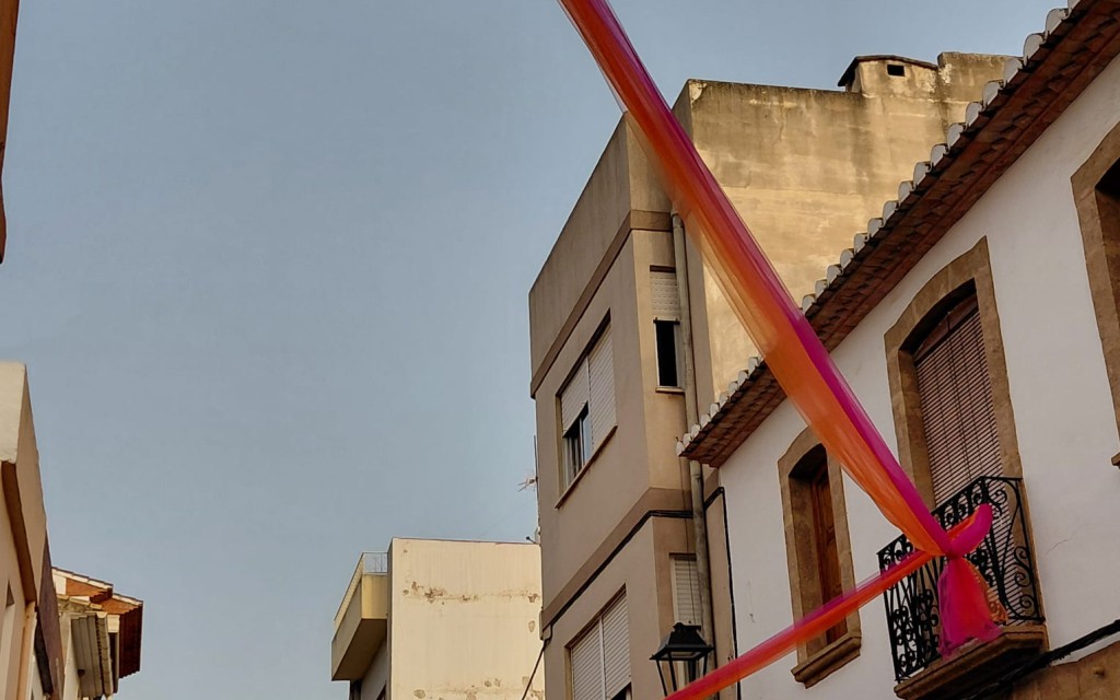 A LES PORTES DE LA FESTA, ADORNANT CARRER. ENTRE TELES I POMPONS, EL CARRER SIGNES MIRA A L'ERMITA