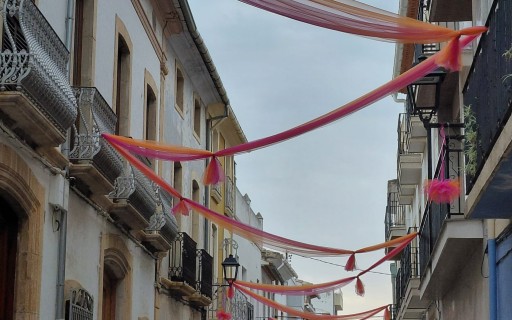 A LES PORTES DE LA FESTA, ADORNANT CARRER. ENTRE TELES I POMPONS, EL CARRER SIGNES MIRA A L'ERMITA